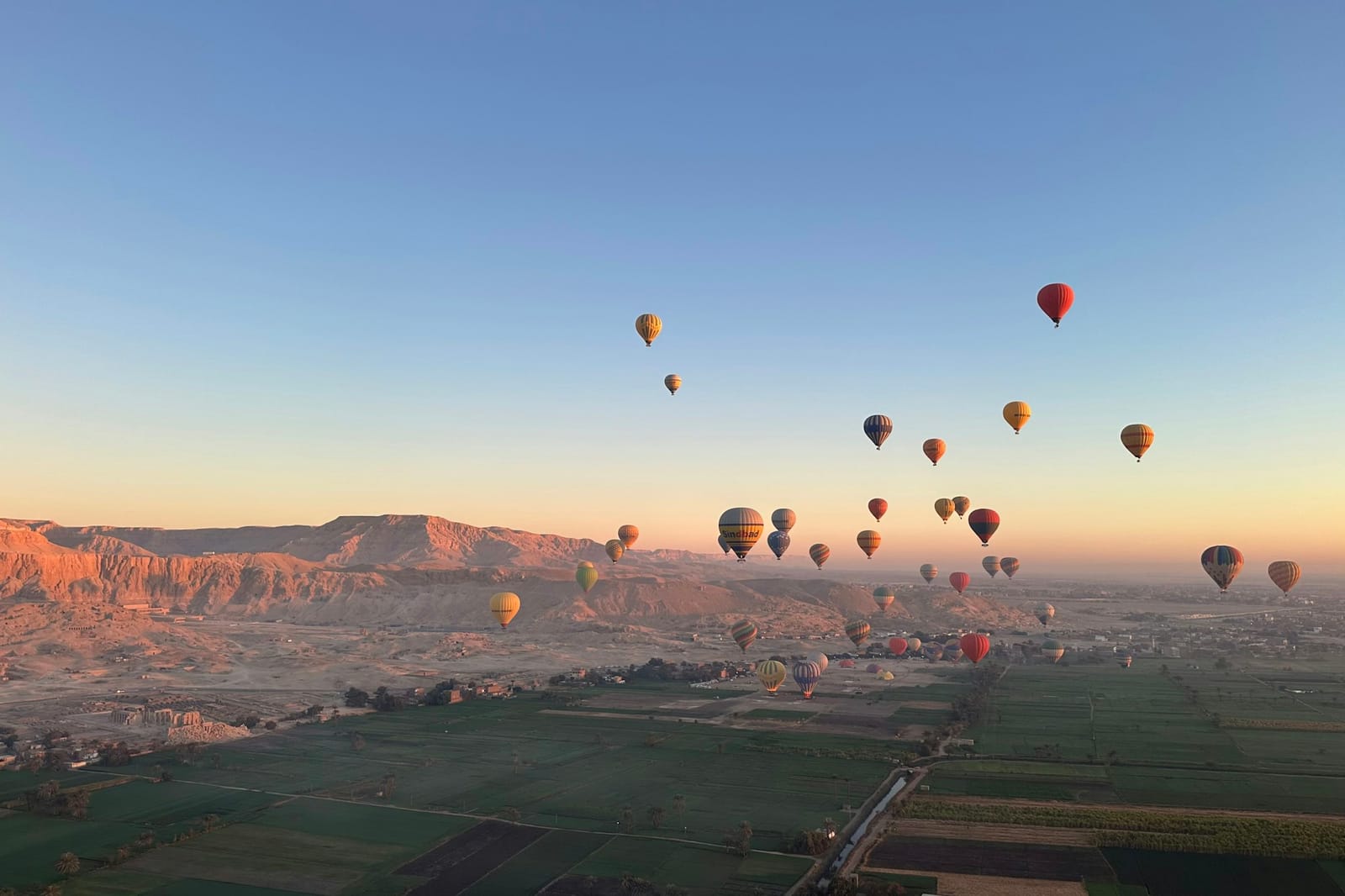 Hot-air balloons of many colours drifting over a wide valley at sunrise.